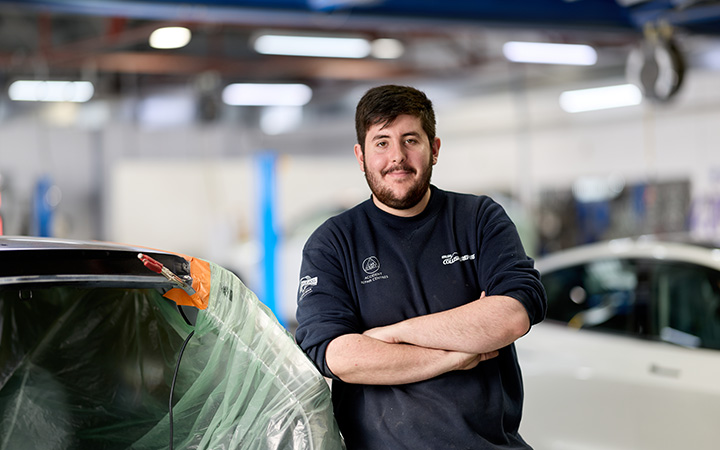 Isaac wearing a branded workshop uniform with arms crossed, positioned beside a car undergoing panel repair.