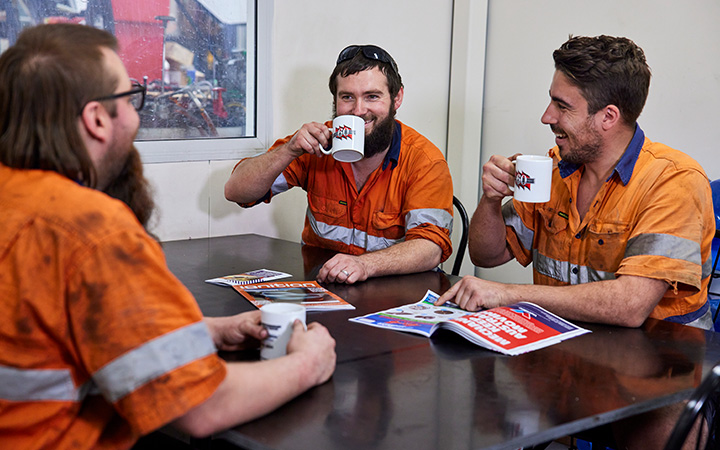 Three mechanics in orange shirts enjoying coffee together at a table, engaged in conversation and laughter.
