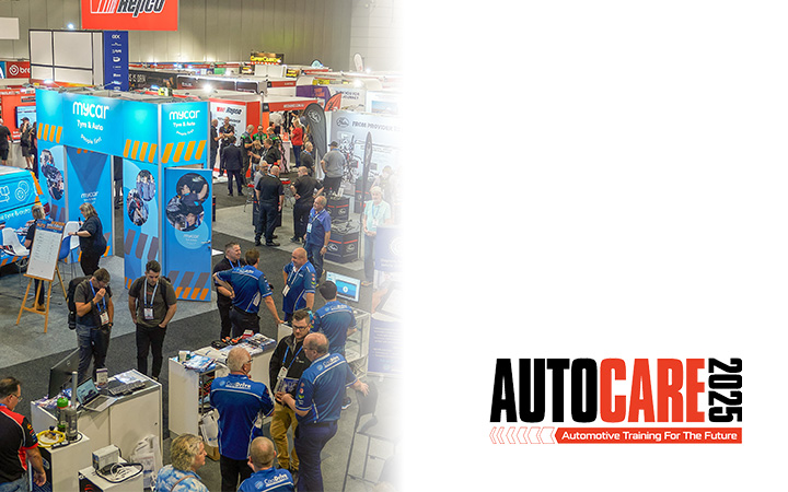 A large crowd of people exploring an auto show (Autocare), with colorful displays and vehicles in the background.