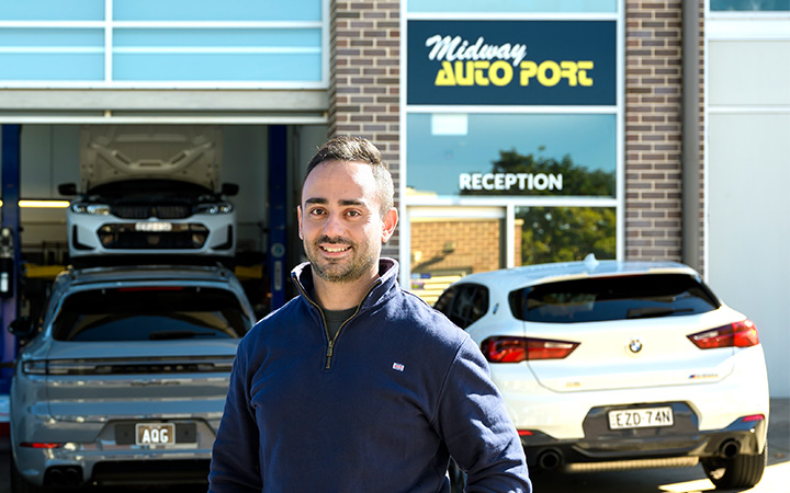 Ross standing outside Midway Autoport workshop in Ryde, with the “Reception” sign visible, cars parked out front and two vehicles on lifts inside the garage.