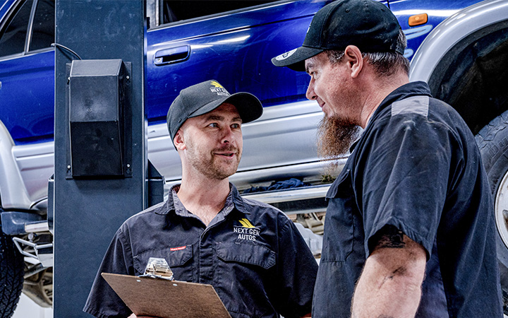 Two automotive technicians in a workshop, engaged in a conversation, with a lifted blue SUV in the background.