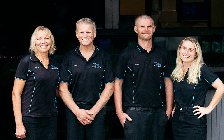 Four Capricorn Members standing together in front of Beenleigh Batteries workshop, wearing black polo shirts with blue accents. From left to right: Steve’s wife, Steve, Darren, and Mikayla, smiling to the camera.