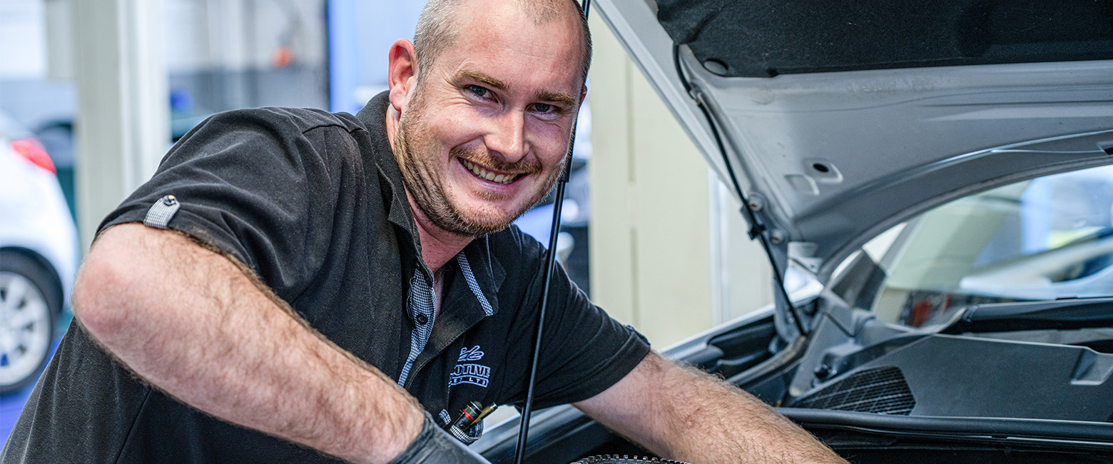 A man smiles while focused on repairing a car, showcasing his enjoyment of automotive work.