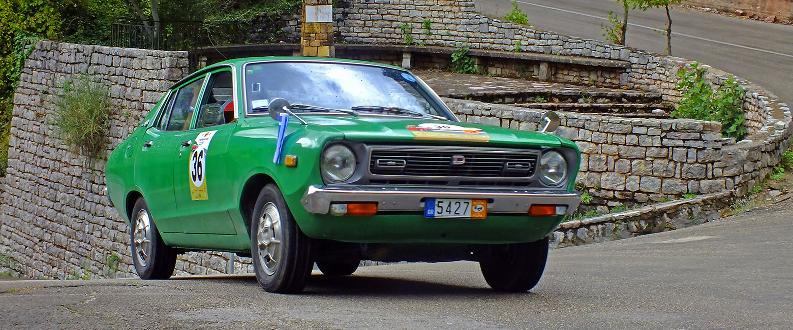 Green Datsun sedan with rally decals driving on a winding road, surrounded by stone walls and greenery.