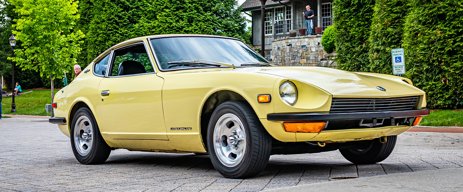 Yellow Datsun 240Z sports car driving on a paved road, with trees and a stone building in the background.