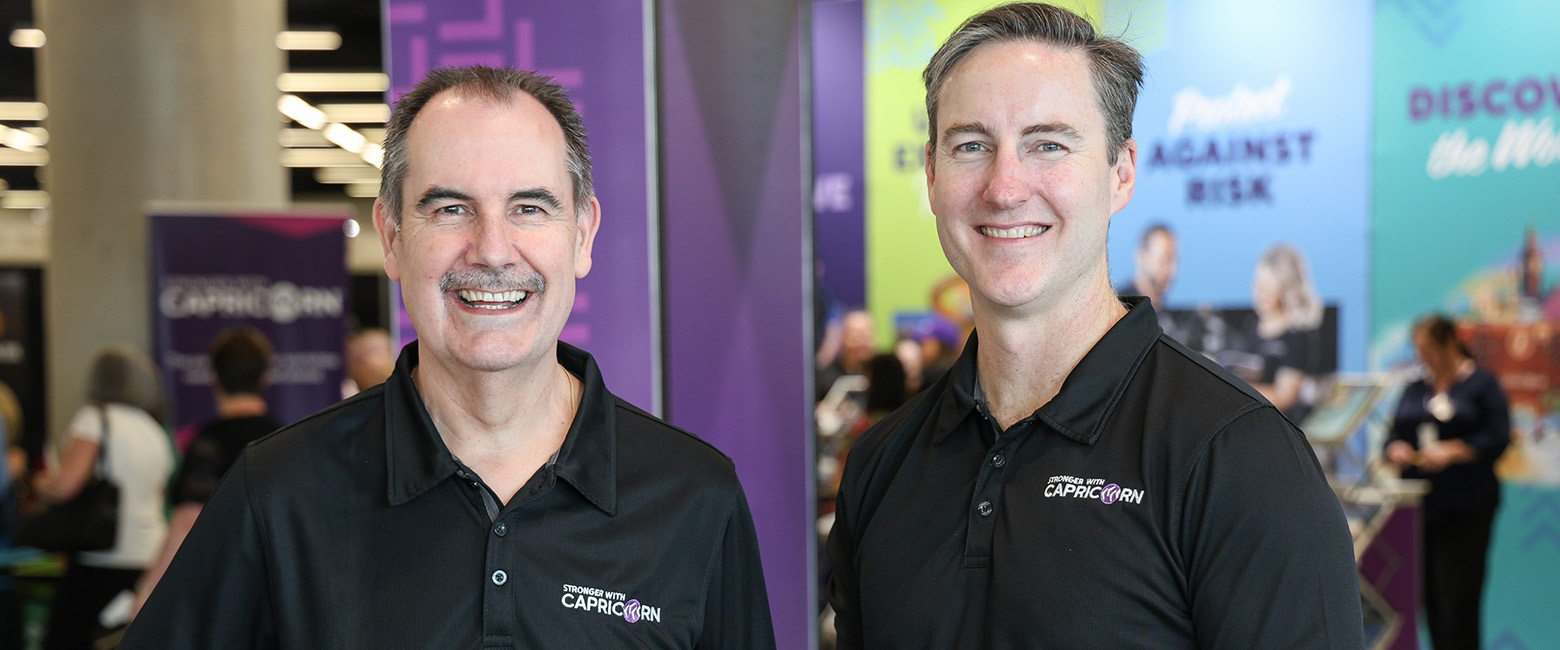 David Fraser, Capricorn Group CEO, and Brad, Capricorn’s incoming Group CEO, both wearing black polos with the Capricorn logo, smiling together at a vibrant event setting, symbolising leadership transition.